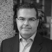 Black and white portrait of a man in a suit and glasses, standing against a textured wall.